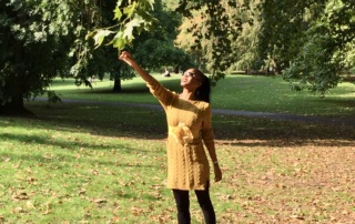 Collette Gee smiling in a mustard sweater dress, reaching toward autumn leaves in a sunny park surrounded by fallen leaves.